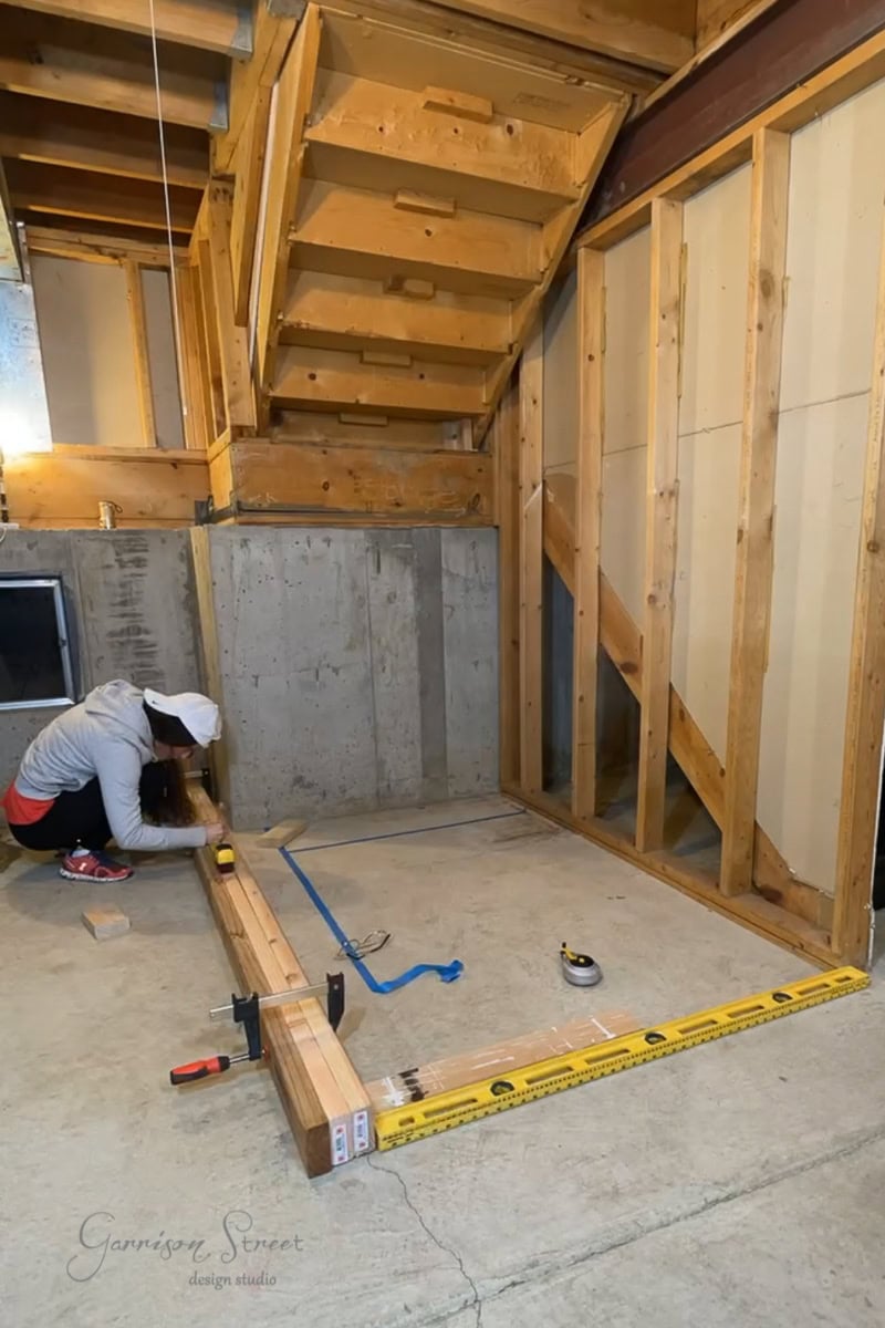 A person in a white cap and red sleeves kneels on a concrete floor, demonstrating how to frame basement walls by measuring and arranging wooden beams. In the background, framed walls and a staircase emerge. Tools and a tape measure rest on the floor.