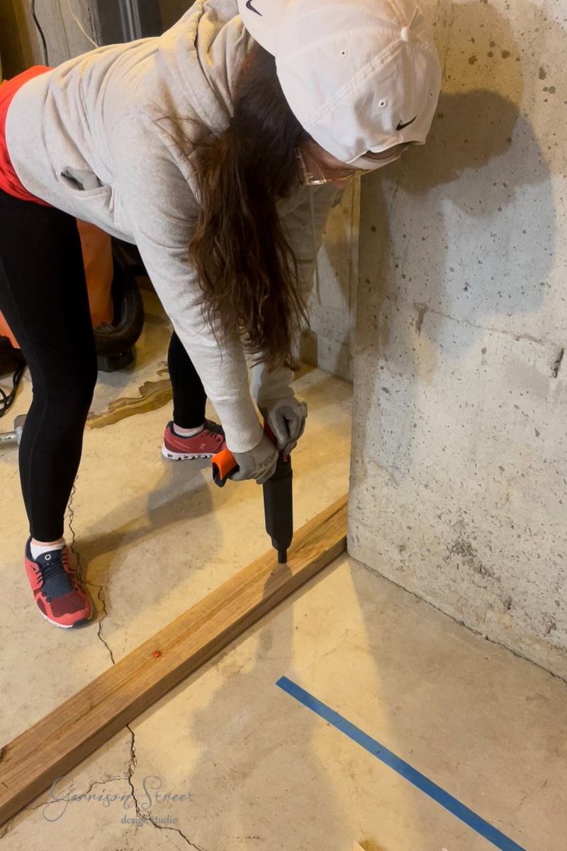 A person wearing a white cap, gray hoodie, black leggings, and red sneakers uses a power tool to work on framing basement walls with a wooden board against a concrete wall. A strip of blue tape is on the floor near the wall.
