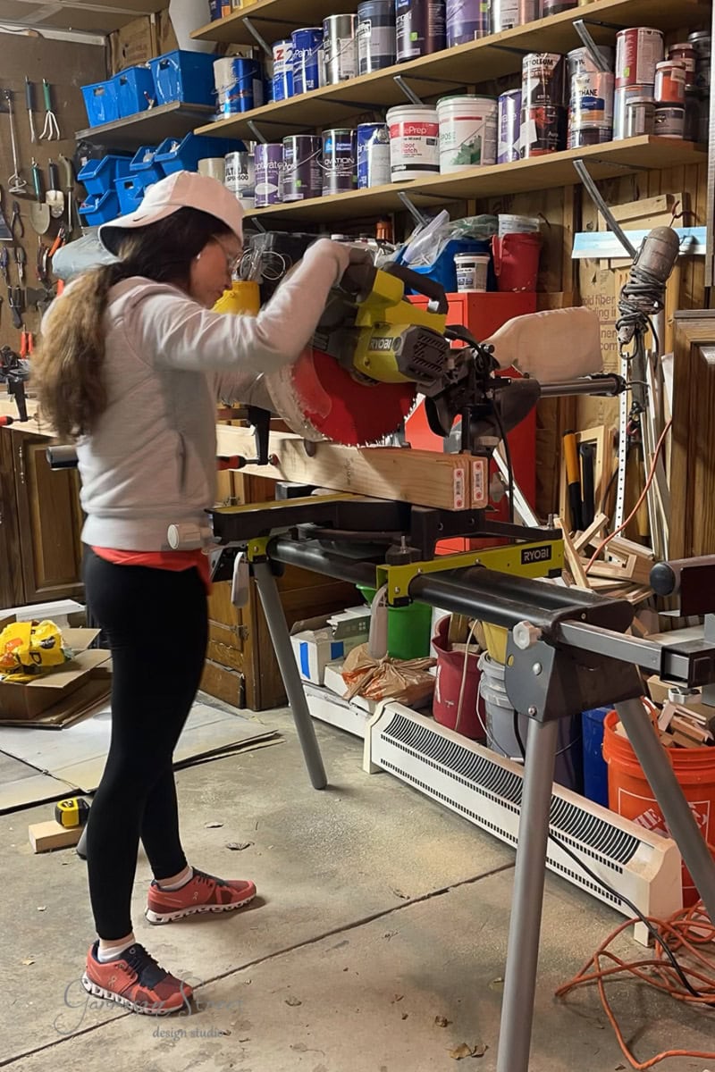 In a cluttered workshop, an individual clad in a cap, long-sleeve shirt, leggings, and sneakers skillfully wields a power saw on wood. Shelves brimming with paint cans and tools surround them as they focus on perfecting techniques crucial for projects like framing basement walls.