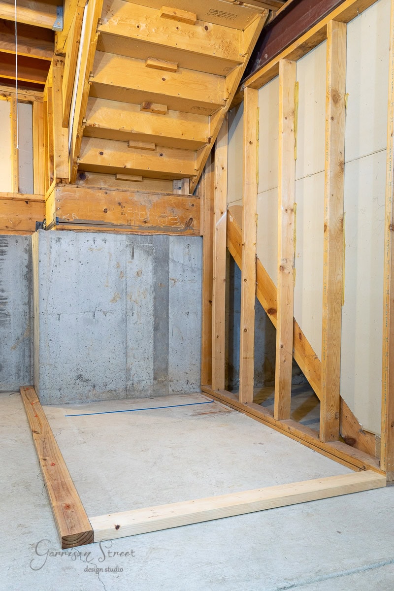 Interior view of an unfinished basement with wooden framing and concrete walls reveals exposed beams and ceiling, complemented by a framed partition wall under a staircase. Natural light pours in, offering a glimpse into how to frame basement walls effectively.