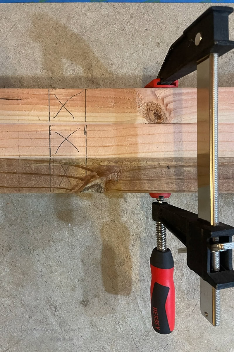 A wooden board, possibly intended for a project like framing basement walls, is clamped to a workbench. Pencil markings and two large X marks adorn its surface. Secured by a red-handled clamp, it rests against a textured gray background, ready for the next step in its transformation.