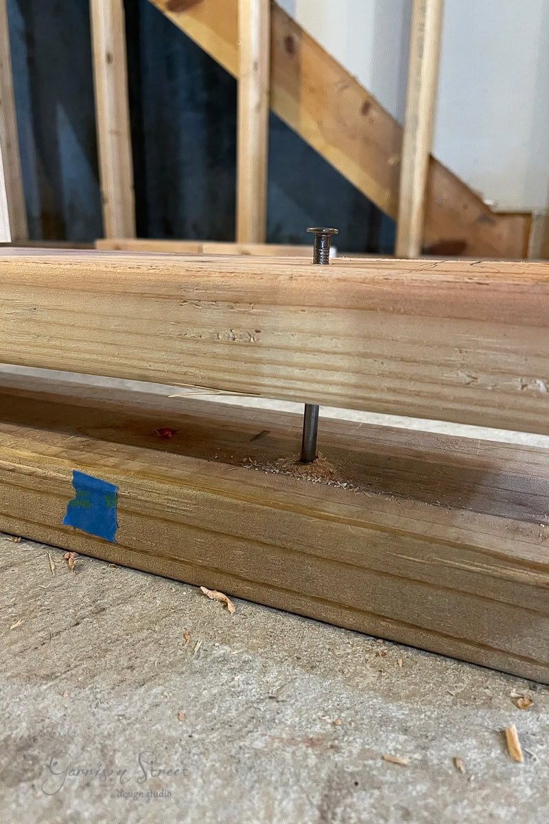 A close-up of a wooden beam with a nail partially driven into it suggests the early stages of learning how to frame basement walls. Resting on a concrete floor with sawdust scattered around, you see the unfinished wall's exposed studs and a piece of blue tape marking progress.