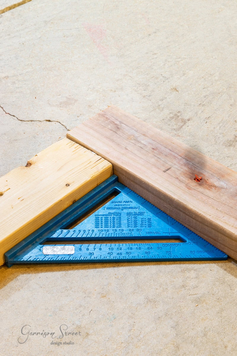 A blue speed square is positioned on a concrete surface, showing how to frame basement walls effectively. Two wooden boards are placed alongside it at a right angle, forming a corner. The board on the right has a small red mark near the edge.