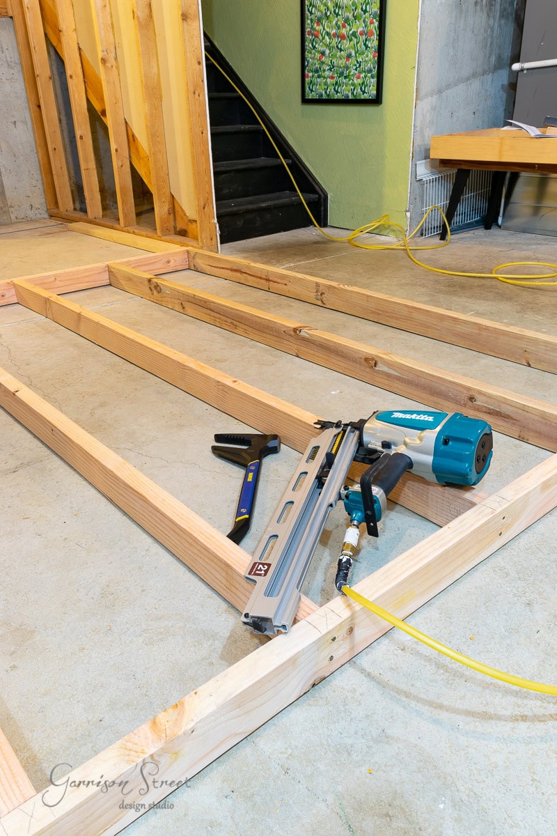 A construction area showcases wooden frames, perhaps illustrating how to frame basement walls. These frames are laid on a concrete floor with a nail gun, hammer, and other tools resting on the wood. In the background, stairs lead up while a framed art piece adds a touch of personality.