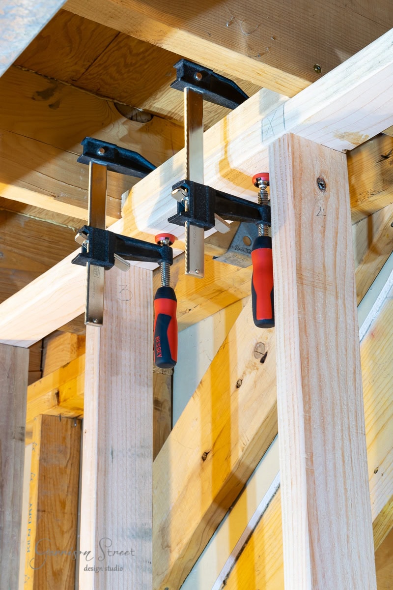 Wooden beams, clamped together with two red-handled metal bar clamps, are part of a larger wooden structure, possibly under construction. The setting mirrors a scene from a guide on how to frame basement walls, with additional wooden elements and framing in the background.