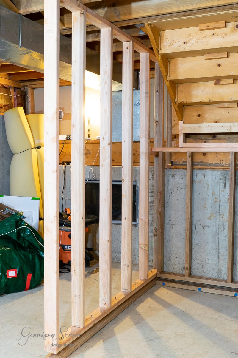 Wooden framing for a wall under construction in a basement area demonstrates how to frame basement walls. The frame consists of vertical wooden studs connected to a horizontal base, situated near a concrete wall and next to a staircase. Various tools and materials are visible around.