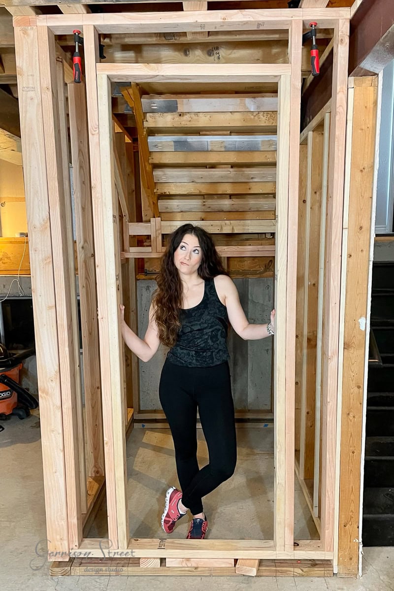 A woman with long dark hair, wearing a black top and pants, poses in a wooden doorway. She stands inside a partially constructed wooden structure in a workshop, demonstrating the essence of how to frame basement walls, with tools and materials scattered around.