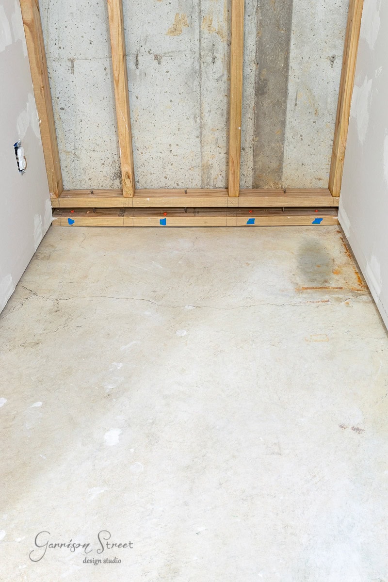 A partially finished room with exposed wooden beams and a concrete wall. The floor, marked in spots, awaits transformation. Drywall panels on the side walls reveal a few visible patches, hinting at an exciting future makeover.