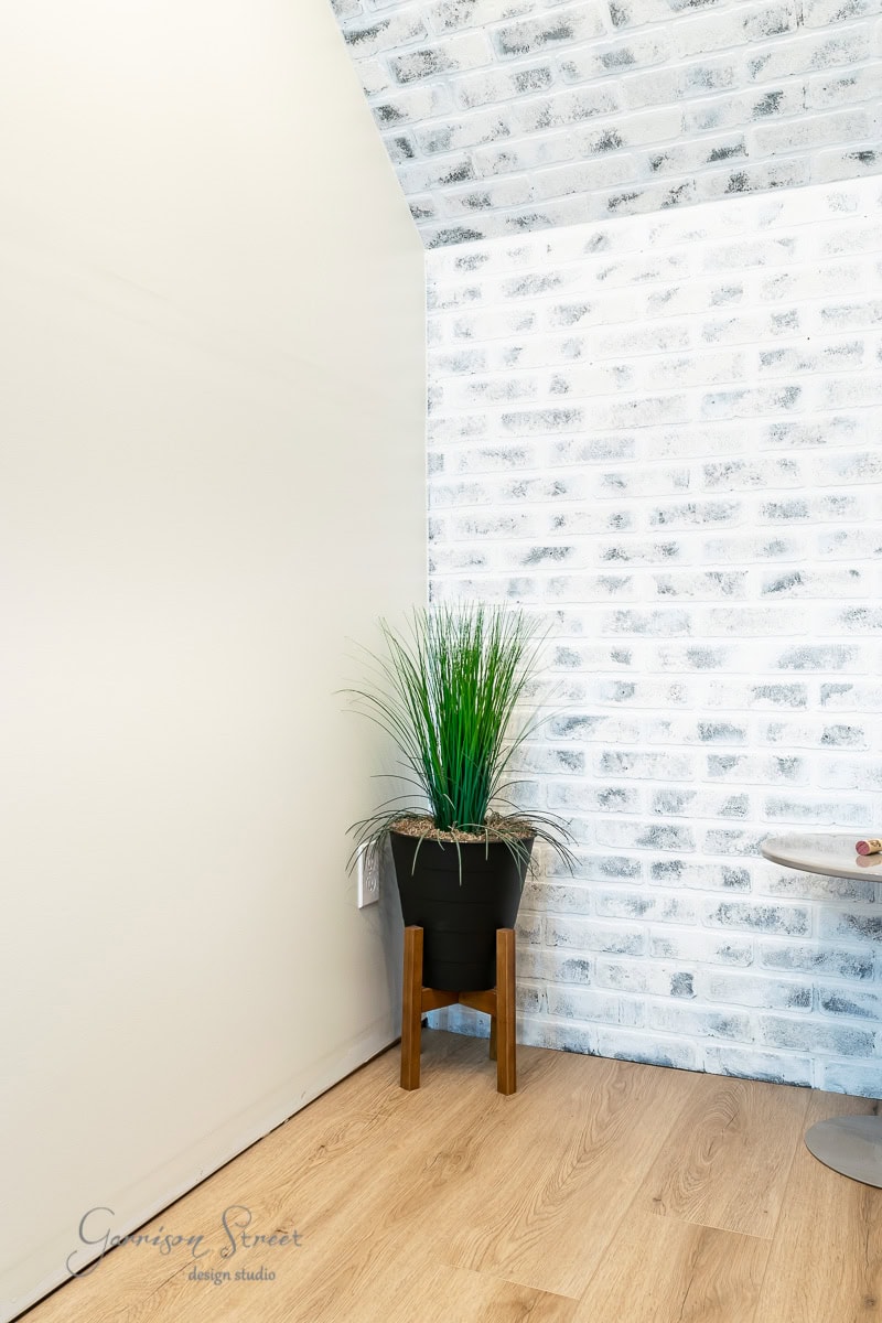 In a modern, minimalist corner of the room, a potted plant with tall grass-like foliage stands elegantly. The whitewashed brick wall contrasts with the light wood flooring. A small table is partially visible on the right.