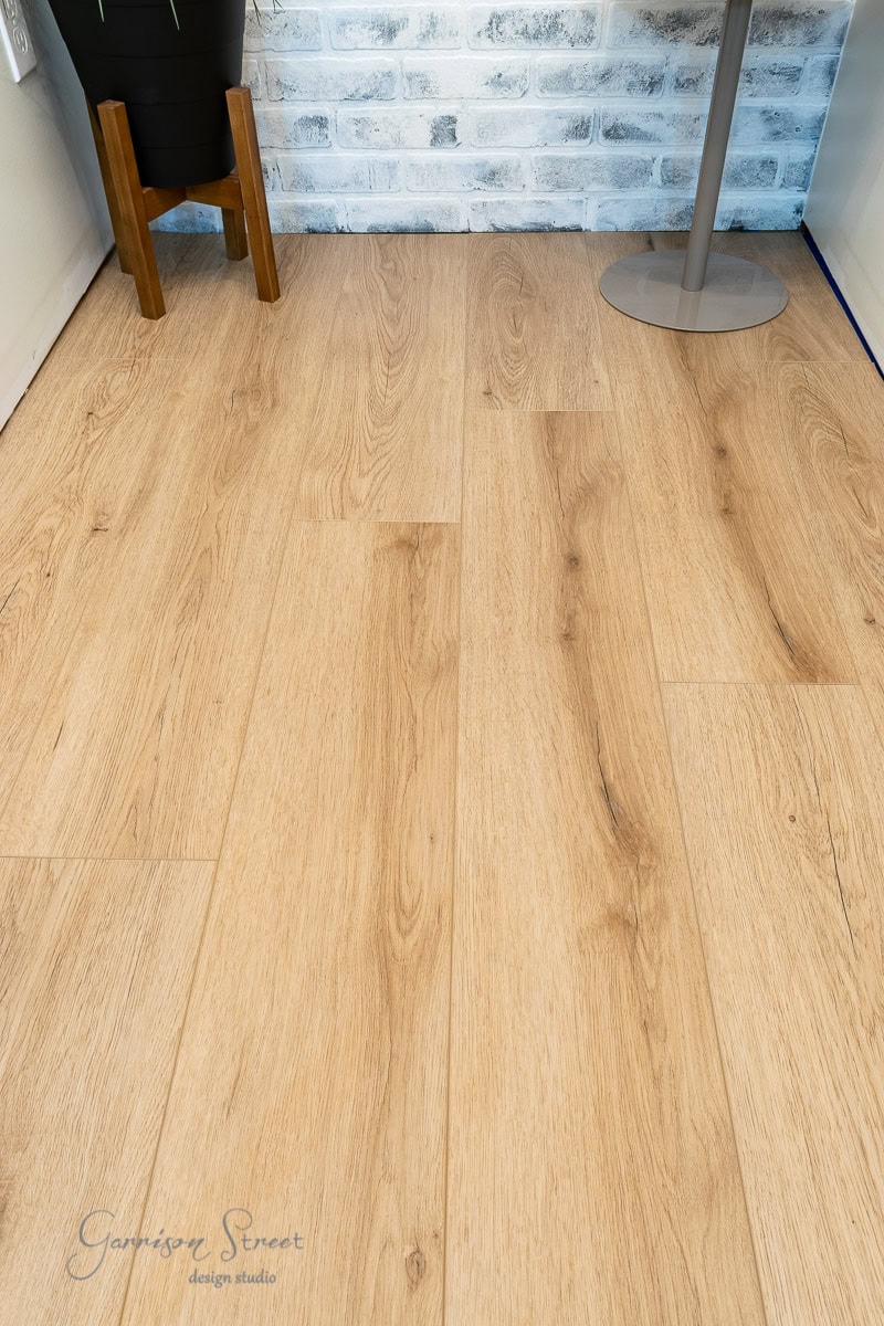 Wooden laminate flooring with a light oak finish covers the basement. It meets a whitewashed brick wall, where a planter and a table with a slender metal base add to the modern and minimalistic decor.