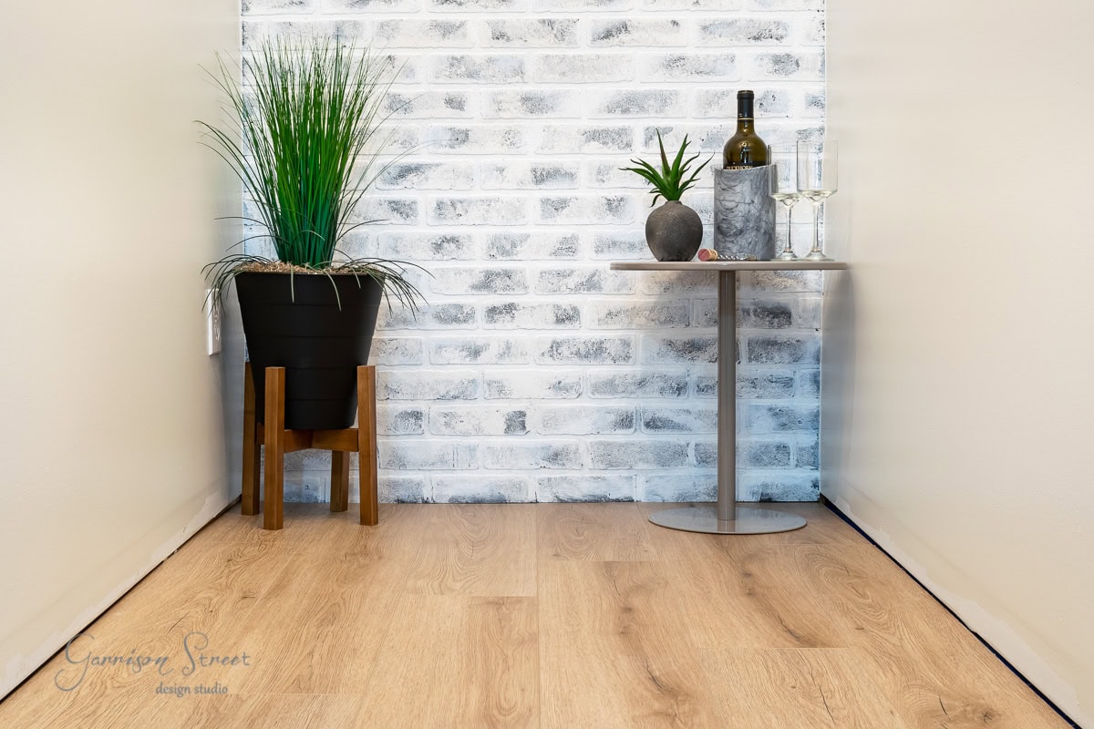 Nestled in a cozy corner with basement flooring options over concrete is a potted plant on a wooden stand. A round table holds a vase, wine bottle, two glasses, and a cut lime. The backdrop of whitewashed brick walls complements the light-colored wood floor beautifully.