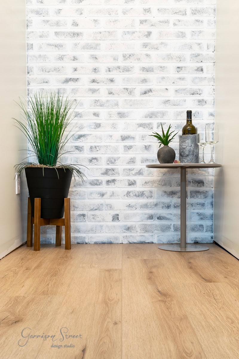 A small, minimalistic corner with light wooden flooring over concrete and a white brick wall. A potted plant stands on the left, and a small table on the right holds a plant, a wine bottle, two glasses, and a decorative object.