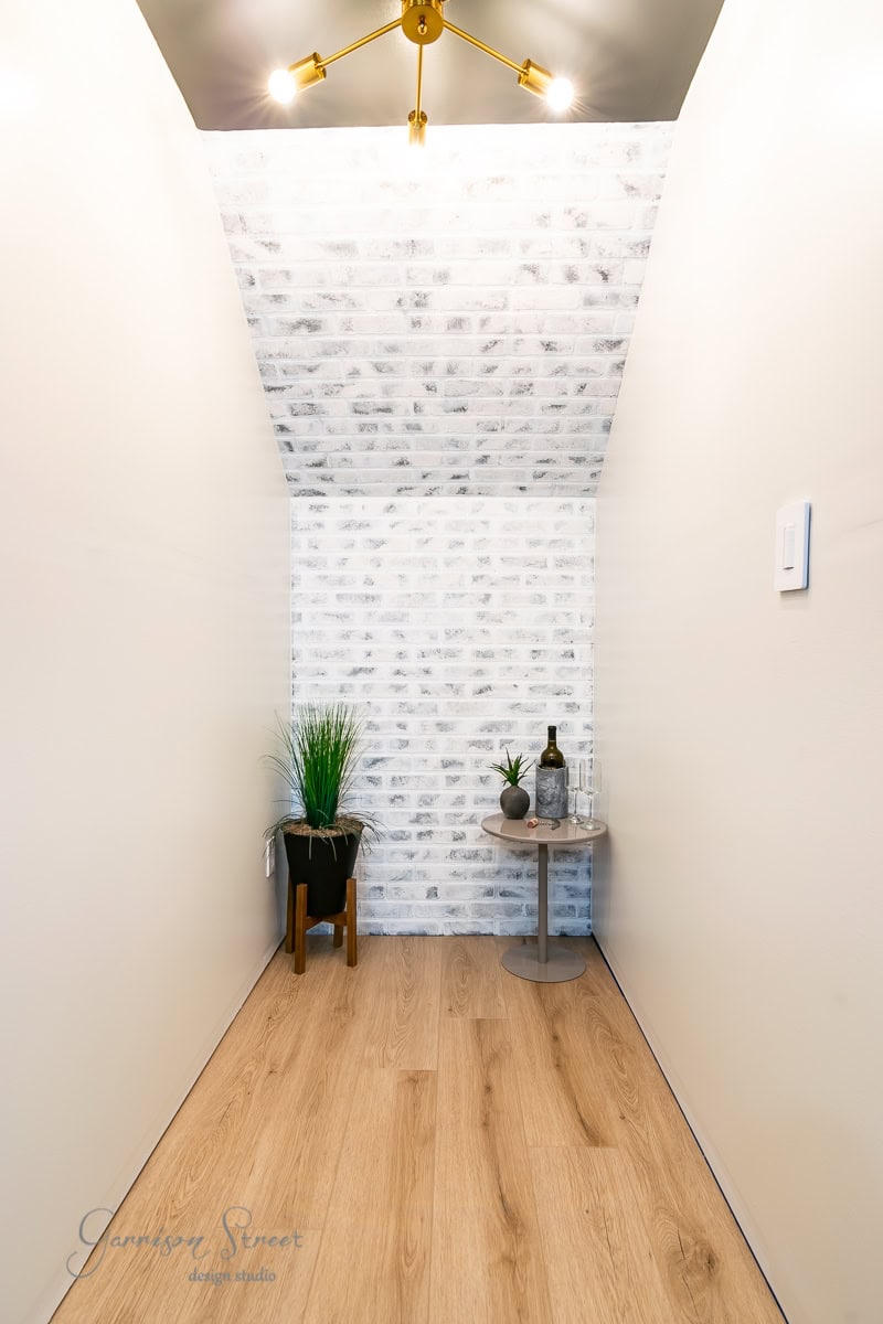 A narrow hallway with light wood flooring showcases a white brick accent wall. A small round table holds a bottle, tray, and decorative items, while a potted plant stands nearby. A modern gold light fixture illuminates the space warmly.