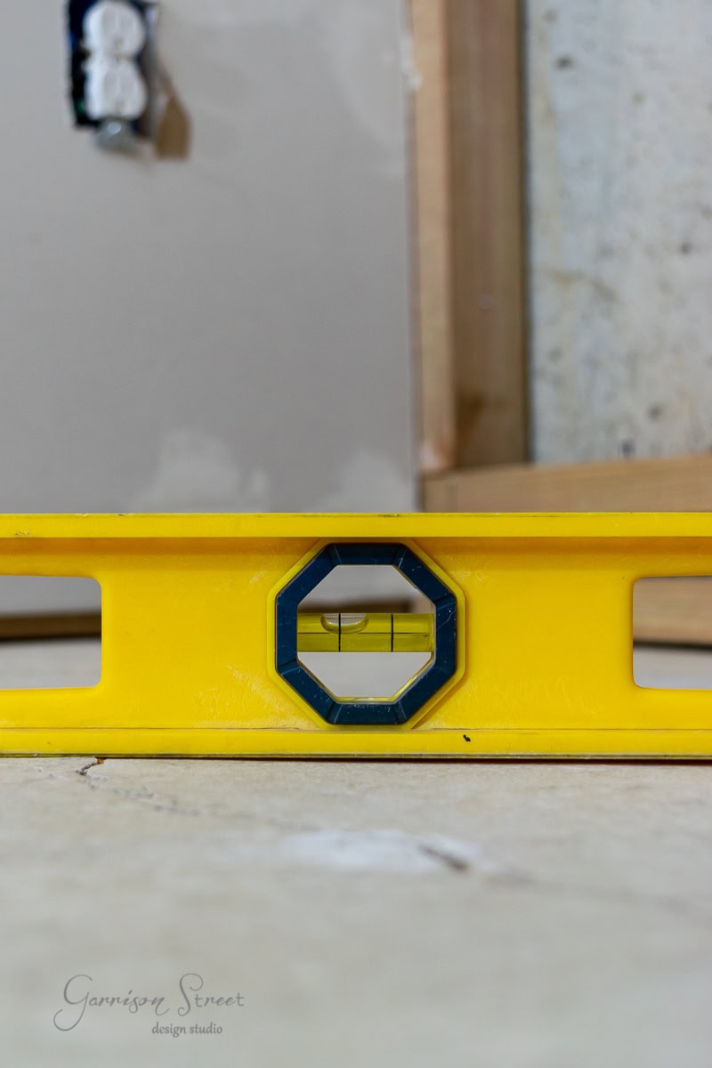 A yellow spirit level rests on a concrete floor, perfectly leveling as the bubble centers itself. In the background, a partially constructed wall with exposed wood framing is visible.