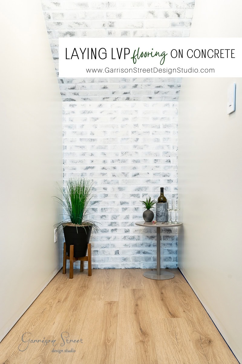 A narrow hallway features light wood flooring. Whitewashed brick walls add charm, while a small round table with a plant and bottles sits on the right. A larger potted plant stands on the left, all under the text "Laying LVP flooring on concrete.