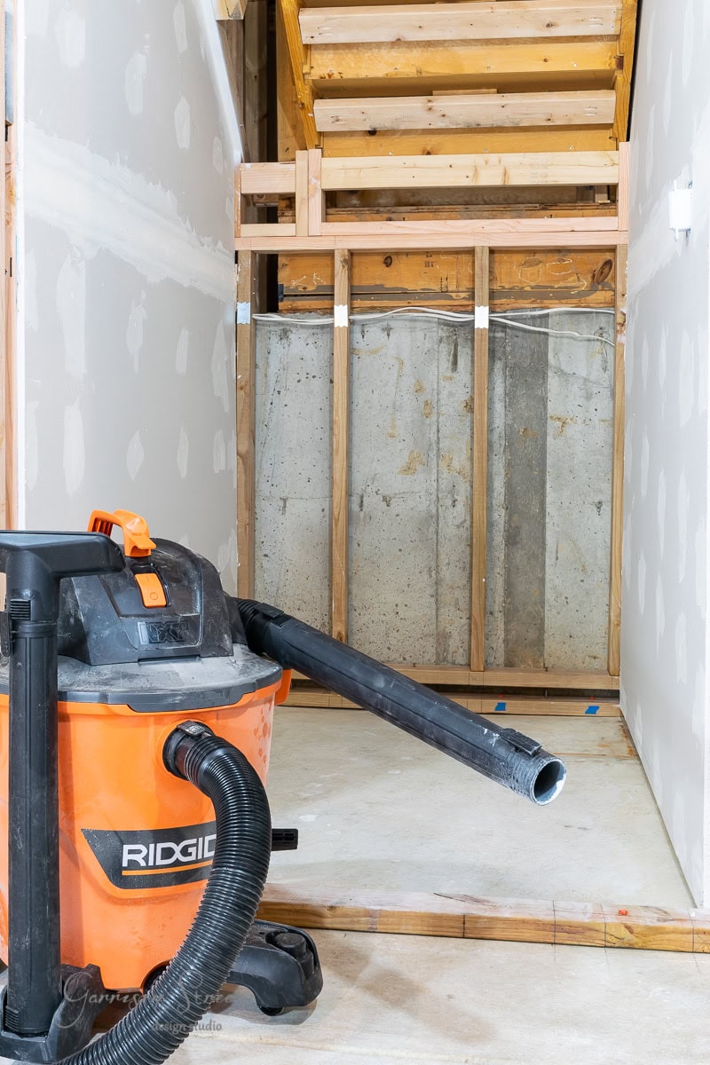 An orange and black Ridgid wet/dry vacuum stands in the foreground of an unfinished under-stair storage area. The space has drywall partially installed and exposed wooden framing.
