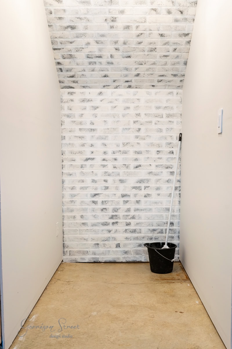 A small, empty nook features a brick-patterned wall at the back and white walls on the sides. In one corner, a mop and black bucket rest quietly on the concrete floor.
