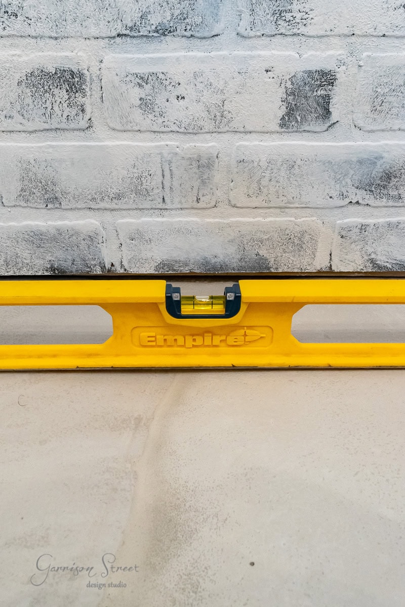 A yellow spirit level rests on a concrete surface against a light-colored brick wall. The level is centered with its bubble visible, ensuring balance.