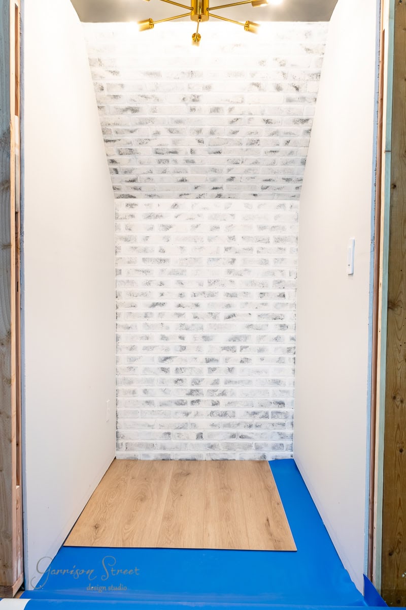 A small, partially renovated closet with white painted brick walls, pristine side walls, a golden ceiling light fixture, blue painter's tape along the base, and a light wood floor.