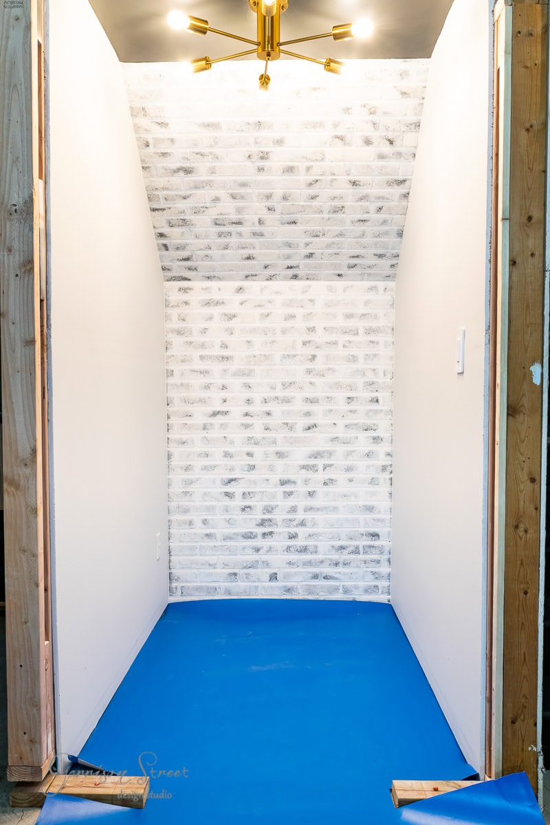 Closet space under construction with white brick back wall, white side walls, and blue plastic-covered floor. A brass chandelier with multiple arms hangs from the ceiling, while wooden framing is visible at the edges.
