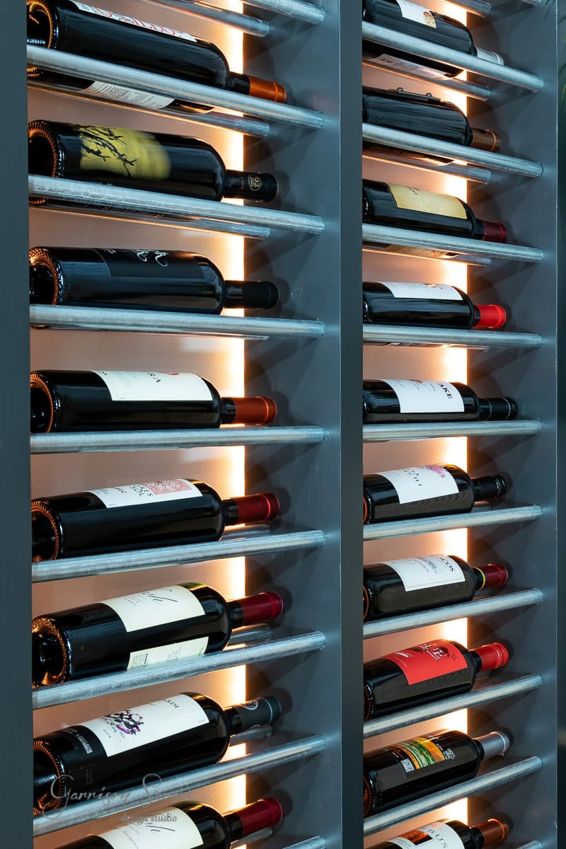 A vertical wine rack boasts an array of assorted bottles. Neatly arranged on horizontal shelves, warm backlighting casts a glow, accentuating the labels and vibrant colors.