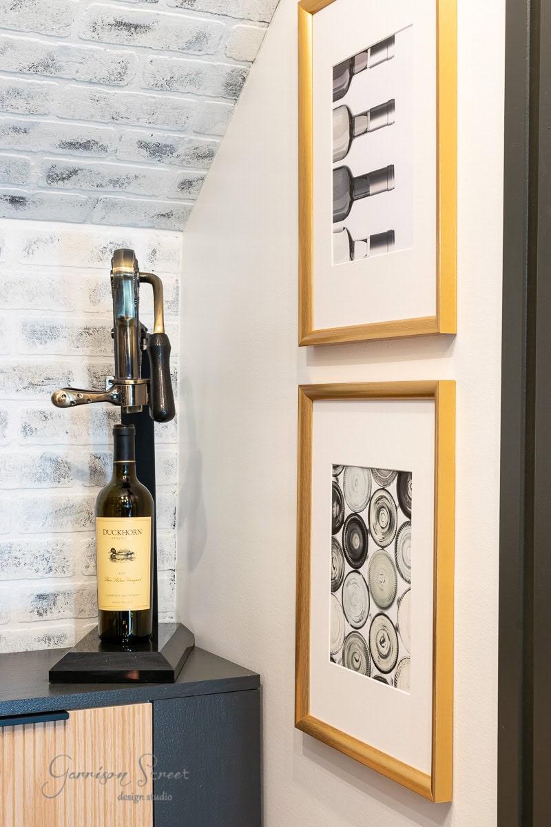 In the corner of the room, a vintage wine opener graces the counter, proudly holding a Duckhorn bottle. Two framed black and white photos of bottle tops and wine bottles adorn the brick wall.