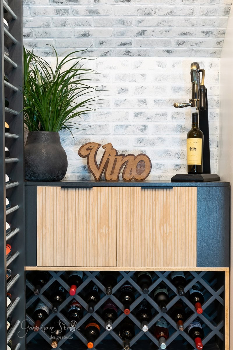 A wine rack filled with bottles is beneath a countertop holding a wooden "Vino" sign, a potted plant, and a corkscrew-adorned bottle of wine, against a white brick wall backdrop.