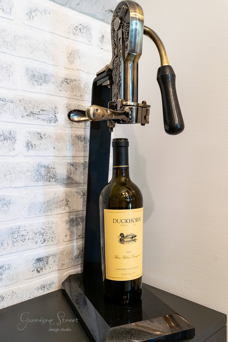 A vintage wine opener stands next to a bottle of Duckhorn Cabernet Sauvignon, set against a white brick wall. The metallic opener with its black handle is mounted on a sleek black base.