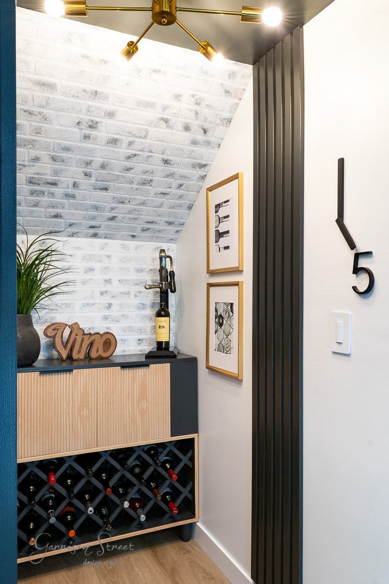 A geometric wine rack filled with bottles, a "Vino" sign, and a potted plant. Art adorns the brick-patterned wall beneath the sloped ceiling, while a modern clock decorates the adjacent space.