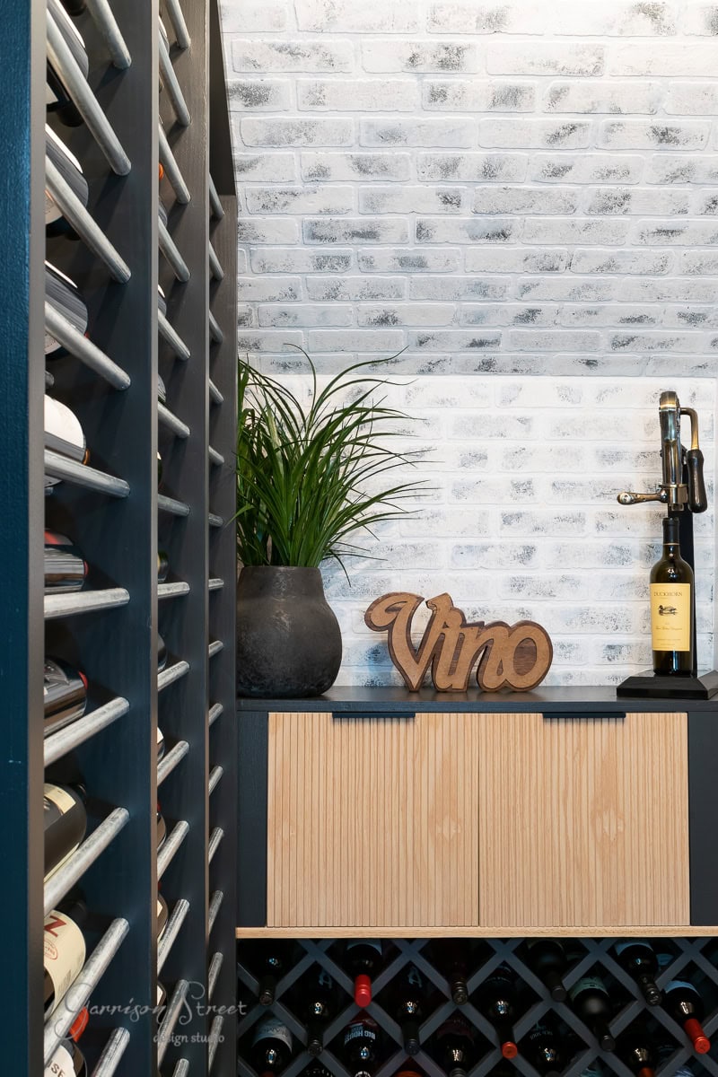 A brick wall backdrop. Wine bottles rest in racks on the left and bottom. A potted plant and a decorative "Vino" sign sit on a shelf beside a wine bottle and corkscrew.