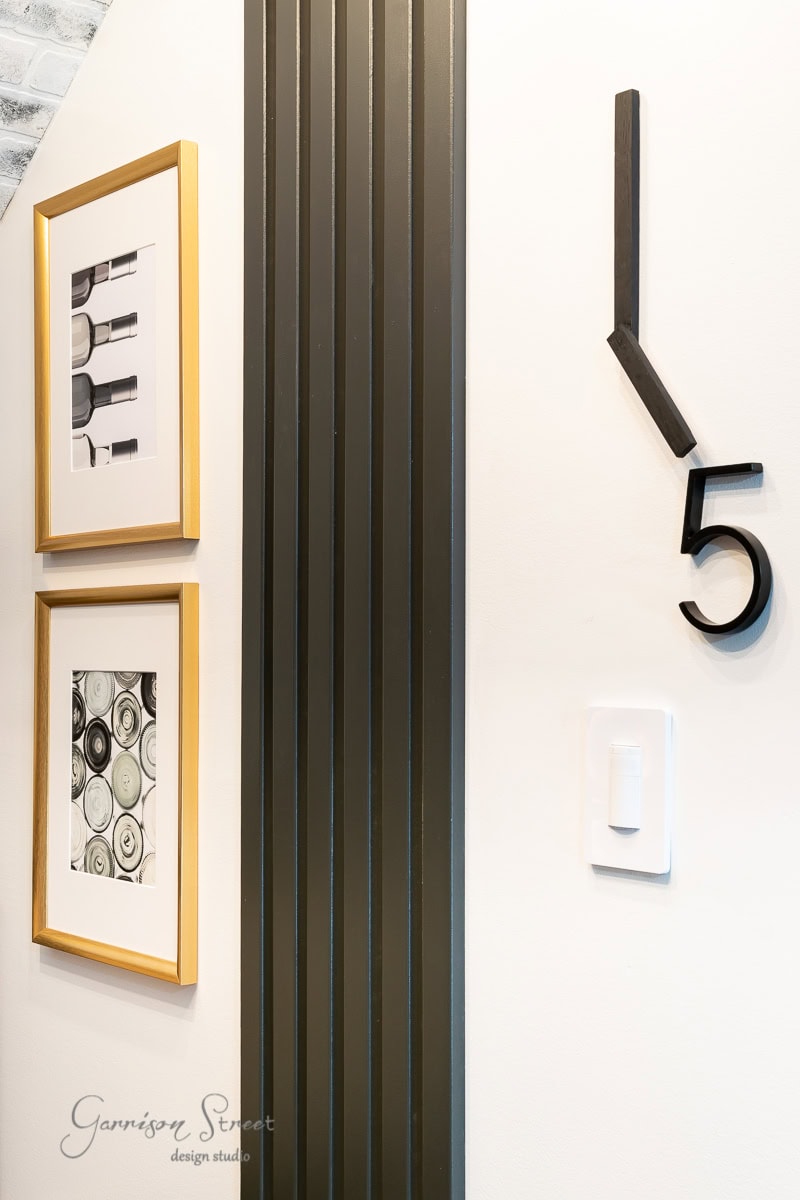 A modern interior wall with artistic details: two framed geometric art pieces with gold frames to the left, a vertical black striped accent in the center, and a minimalist wall clock showing 5 o'clock to the right.