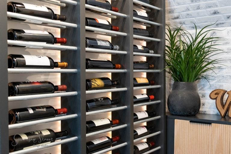 A DIY basement wine cellar features a wine rack brimming with various bottles against a brick wall. To the right, a large gray planter holds a green leafy plant. A wooden cabinet and decorative item are partially visible, creating a well-lit and orderly scene.