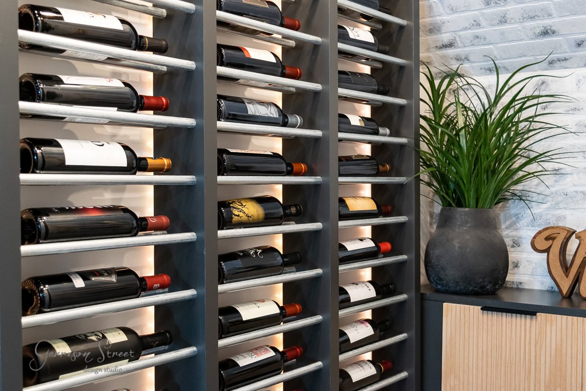 A DIY basement wine cellar features a wine rack brimming with various bottles against a brick wall. To the right, a large gray planter holds a green leafy plant. A wooden cabinet and decorative item are partially visible, creating a well-lit and orderly scene.