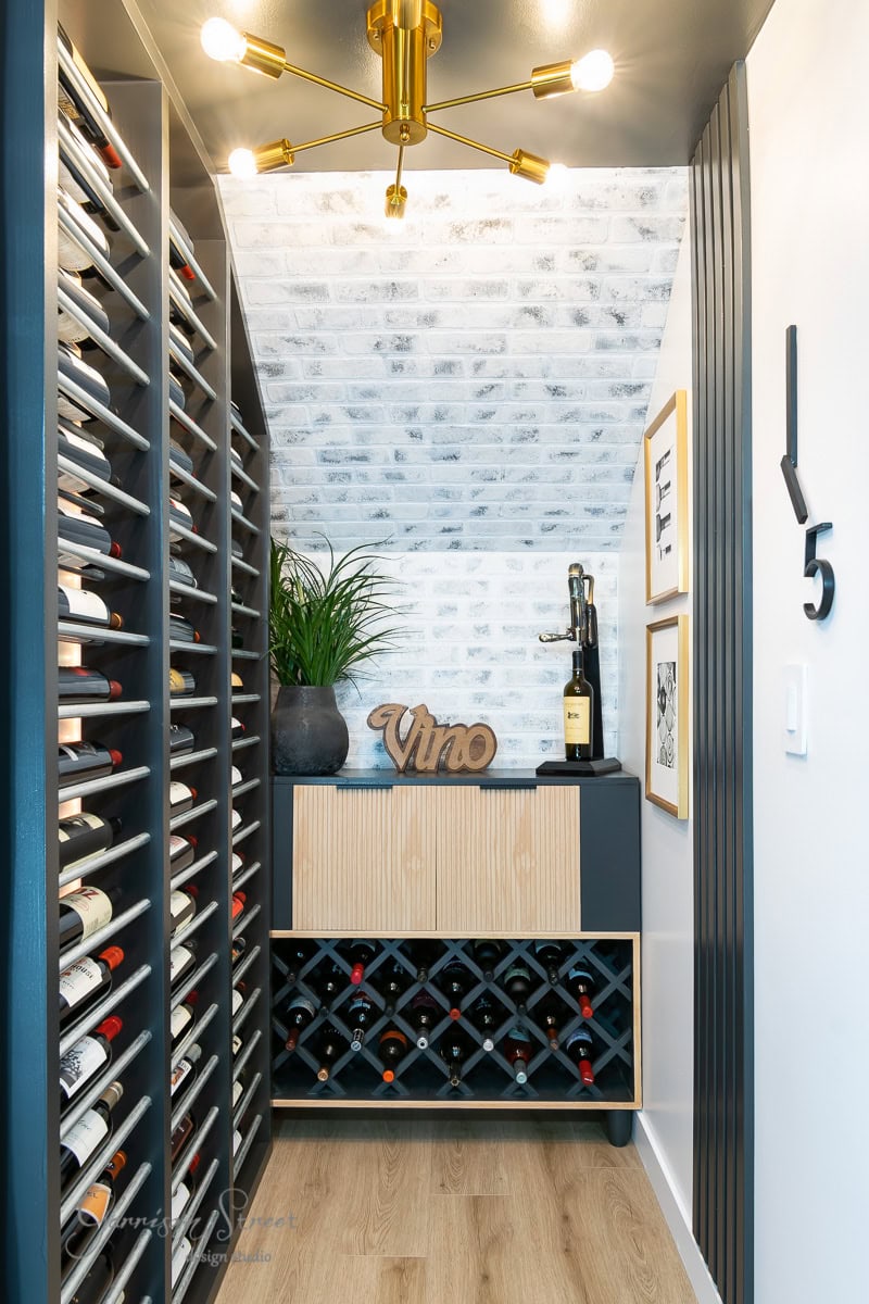 Wall-mounted racks brim with bottles. A wooden cabinet adorned with a "Vino" sign, a plant, and wine tools sits below. From the ceiling hangs a stylish light fixture, casting a glow over walls embellished with artwork.