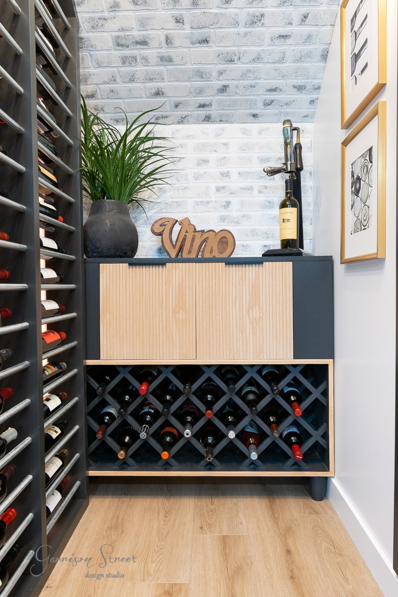 A blue cabinet with a grid wine rack filled with bottles, a wooden "Vino" sign, and a plant. On top sit corkscrew and glasses. The textured wall hosts framed art.