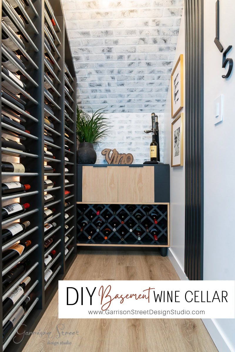 Two racks of wine bottles sit on the left. The back wall displays a small cabinet adorned with a decorative "Vino" sign and framed pictures. A wooden floor complements the white brick-textured wall.