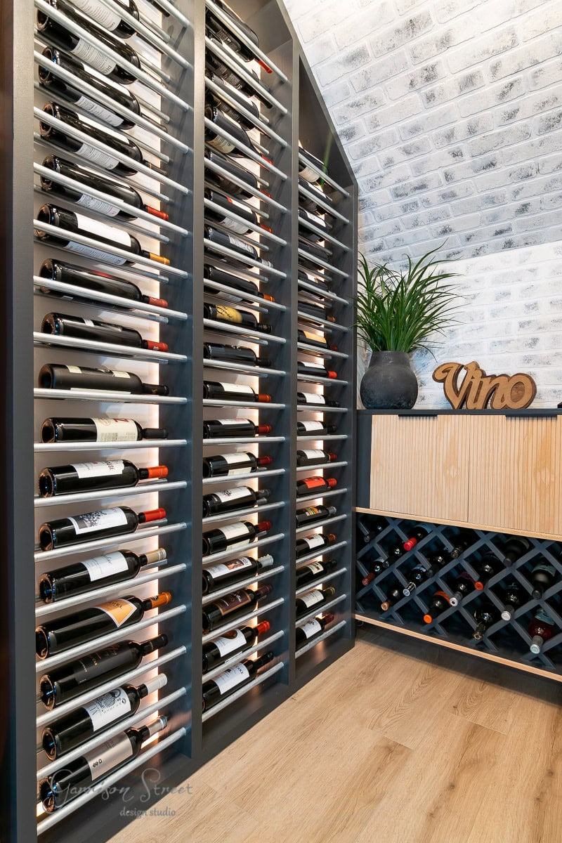 A floor-to-ceiling rack teeming with wine bottles. A small cabinet, holding more bottles and a decorative plant, rests against the white brick wall. The wooden "Vino" sign is displayed on the cabinet.