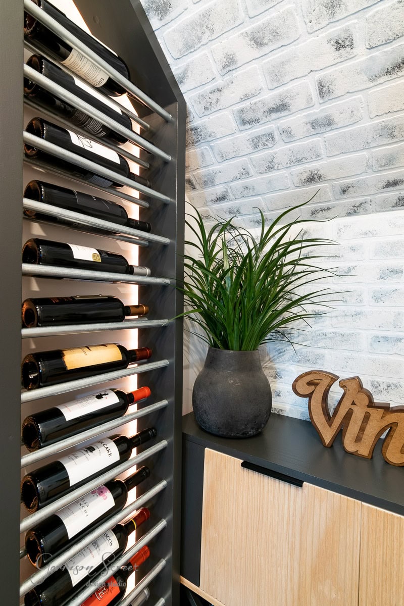 A vertical rack with wine bottles, a black pot with green foliage, and a wooden cabinet. The white-painted brick wall adds rustic charm, and a wooden "Vin" sign completes the space.