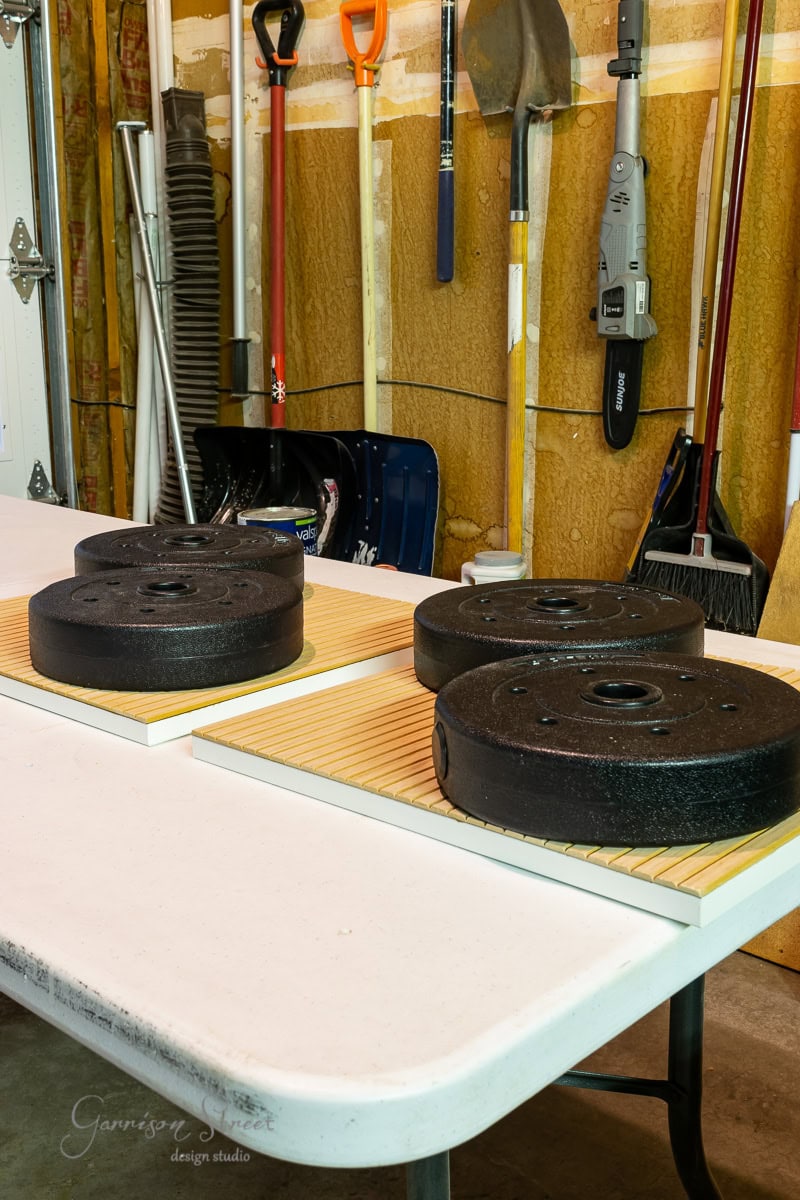 In a garage filled with tools and equipment, weights rest on a table supported by boards. Nearby, shovels and brooms lean against a plywood wall. It's an unexpected setting for an IKEA wine bar cabinet hack, blending practicality with creativity in the heart of this workspace.