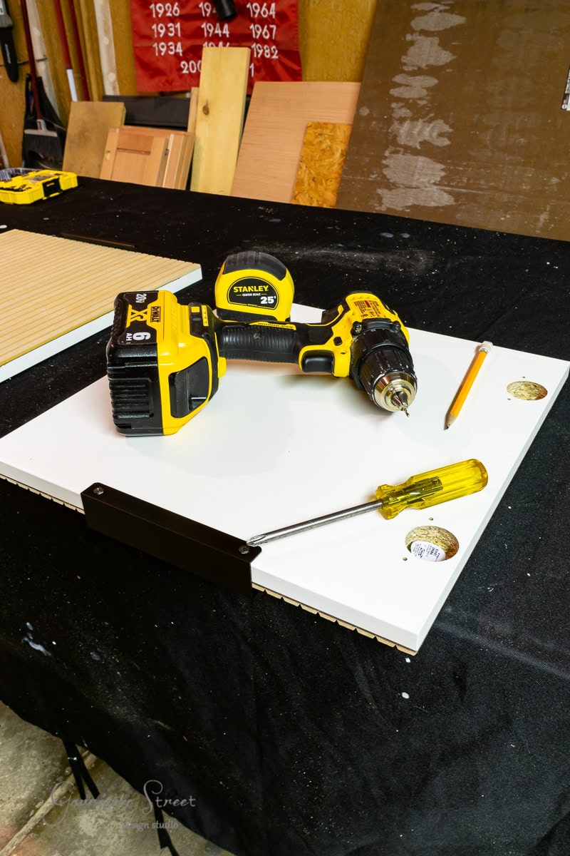 A power drill, screwdriver, and pencil lie on a white panel against a black tablecloth, ready for the ultimate IKEA wine bar cabinet hack. A yellow tape measure is nearby. Posters with years from 1922 to 1970 are visible in the background, adding a vintage touch to this DIY project.