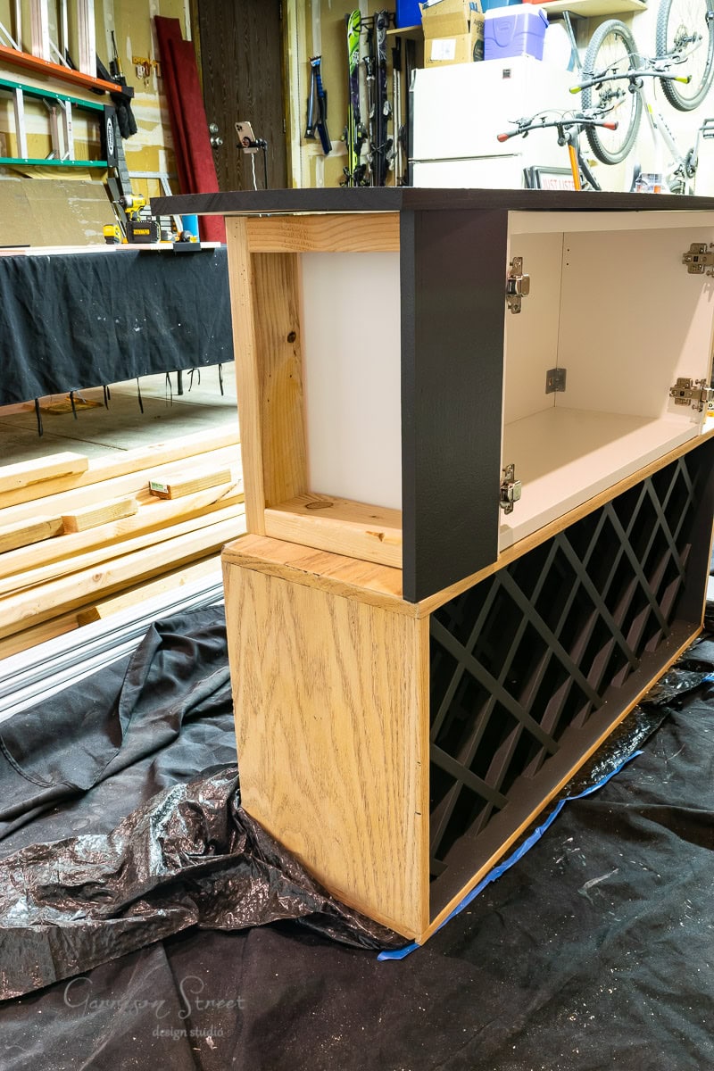 A partially assembled wooden cabinet with a wine rack section sits on a black tarp in the workshop, showcasing the beginnings of an IKEA wine bar hack. Wooden boards and construction materials fill the background, while a bicycle hangs casually on the wall.
