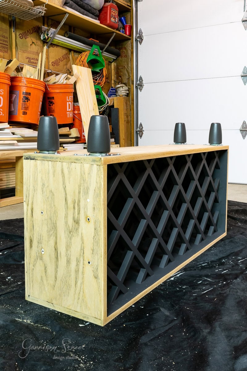 A wooden wine rack, part of an IKEA wine bar cabinet hack, lies upside down on a black tarp in a workshop. It features black lattice for bottles and four inverted legs, with various tools and materials scattered in the background.
