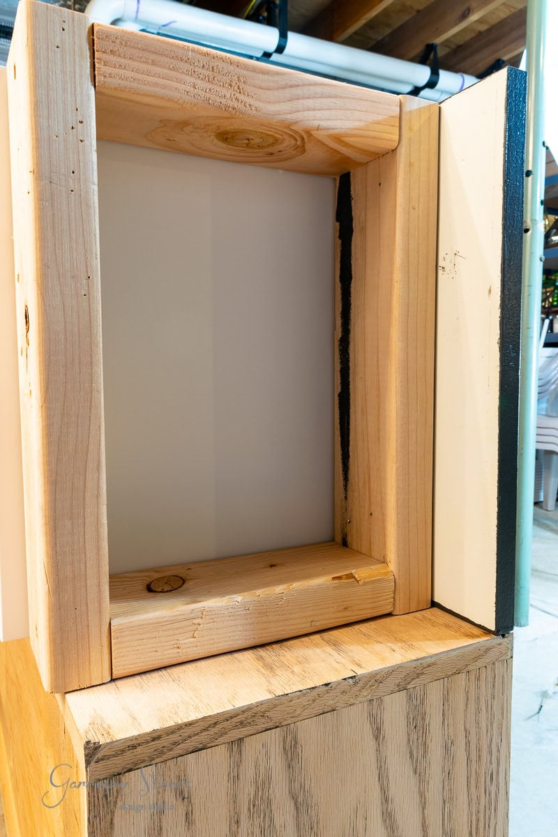 A close-up view of an empty wooden frame sitting on a wooden surface evokes thoughts of an IKEA Wine Bar Cabinet hack. The unfinished frame showcases the natural wood grain, while the background is softly blurred, with some pipes visible above.
