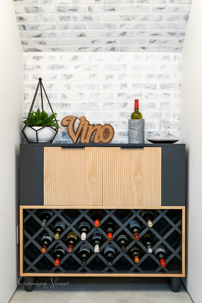 A cozy home wine storage area features an IKEA wine bar cabinet hack, a wine rack stocked with bottles, a small cabinet adorned with a plant, and a charming wooden "vino" sign. An open bottle of wine and two glasses rest nearby, all set against white brick-style wallpaper.