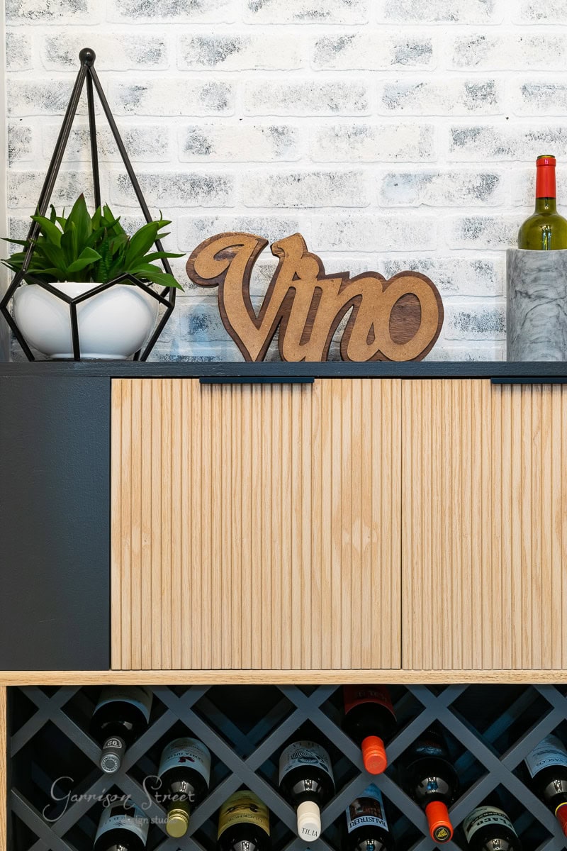 A modern wine display showcases an IKEA Wine Bar Cabinet Hack with a built-in rack for several bottles. Above the sleek cabinet, a wooden "Vino" sign complements a geometric plant holder. The textured white brick wall serves as a charming backdrop.