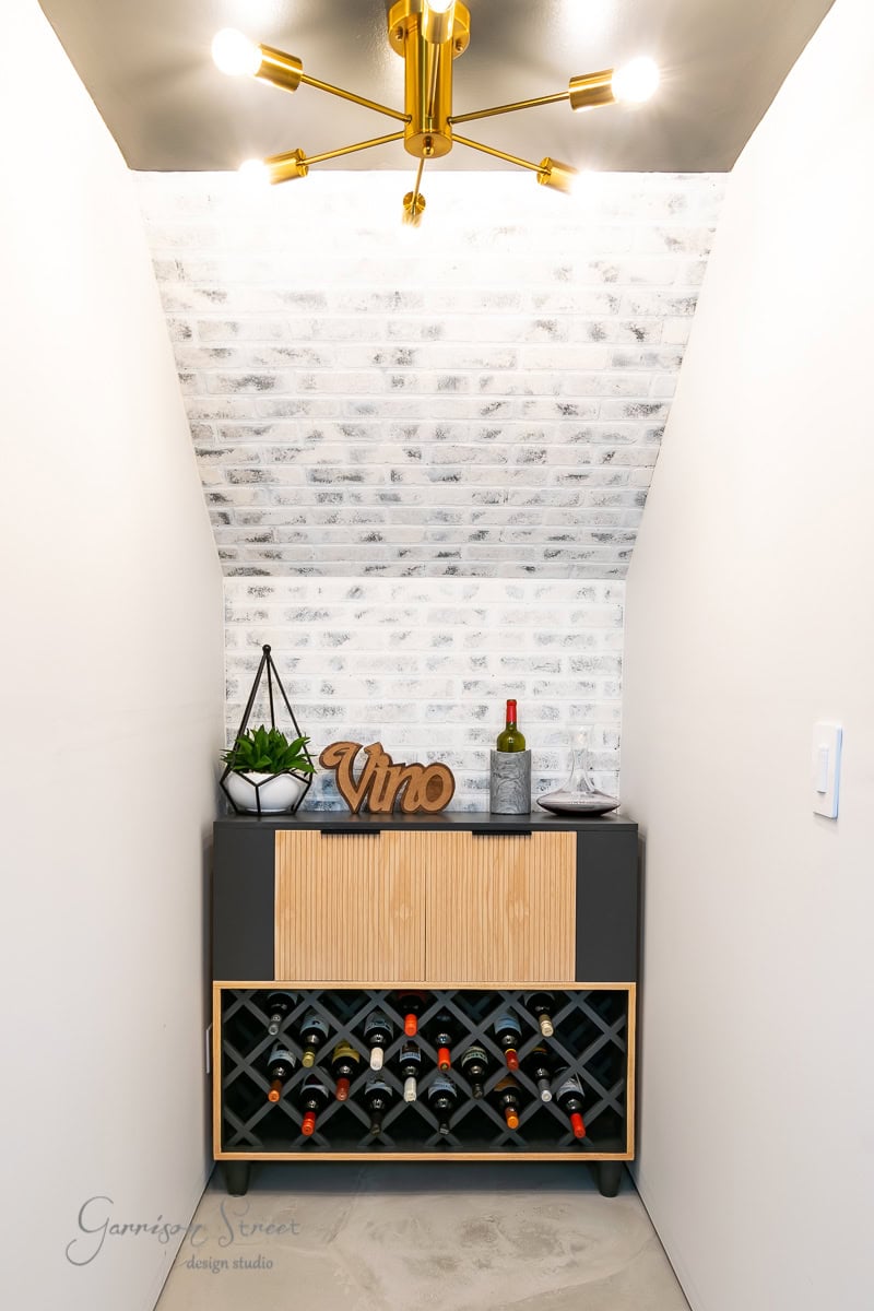 A small wine nook showcases a modern black and wood wine rack, akin to an IKEA Wine Bar Cabinet Hack. It elegantly holds several bottles, adorned with a "Vino" sign and a plant. The white brick wall behind complements the scene, enhanced by the warm glow of a gold light fixture above.