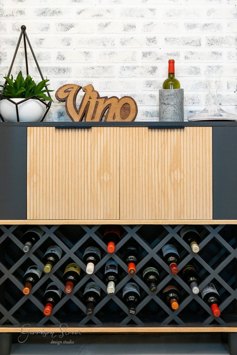 A wine rack with multiple bottles is set against a white brick wall. Above it, an IKEA wine bar cabinet hack features a wooden shelf displaying a potted plant, a sign reading "Vino," and a bottle of wine next to empty glasses.