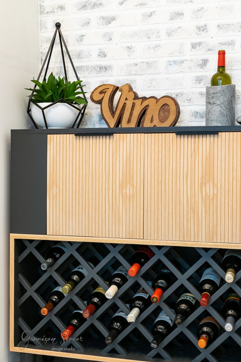 A creative IKEA Wine Bar Cabinet Hack transforms a wooden wine rack filled with various bottles against a white brick wall. Topped with a "Vino" sign and a small potted plant, this setup combines functionality with style.