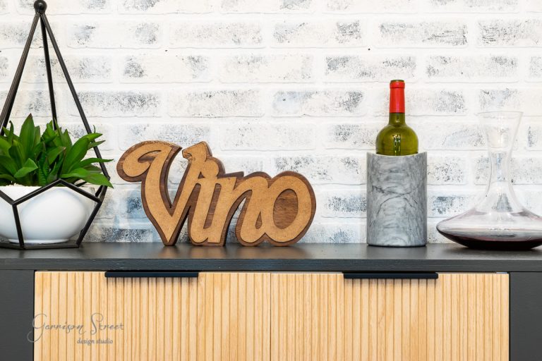 A decorative piece spelling "Vino" rests on a countertop, next to a bottle of wine in a marble cooler, a wine decanter, and a potted plant. The setup feels like an IKEA Wine Bar Cabinet hack against the backdrop of a chic white brick wall.