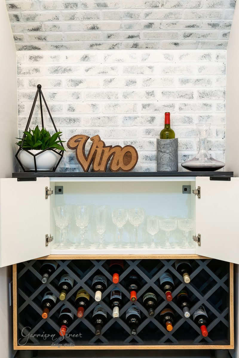 A cozy wine nook with a brick wall highlights an IKEA wine bar cabinet hack, featuring a rack brimming with bottles, a wooden "Vino" sign, and a plant in a geometric pot. Wine glasses are neatly stored behind glass doors, while a bottle and two decanters are artfully on display.